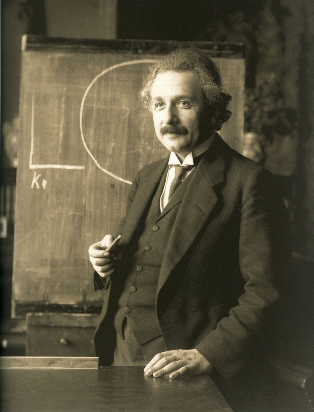 Albert Einstein in a dark suit, standing at a desk in front of a chalkboard, photographed in 1921
