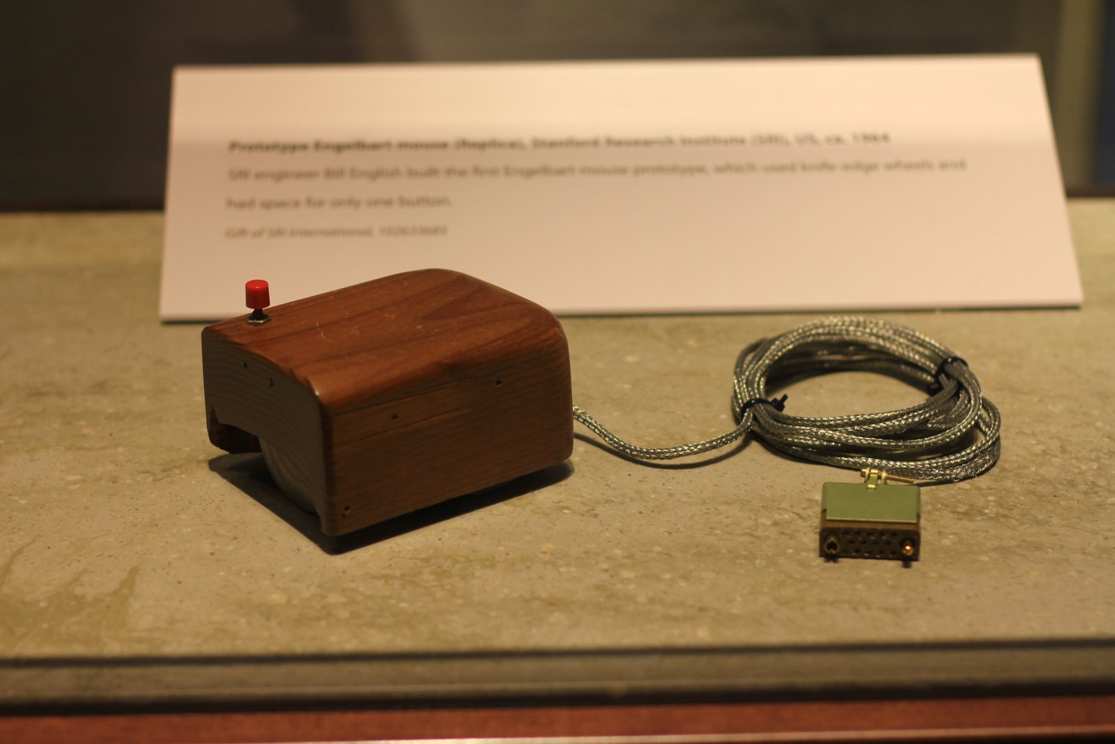 Douglas Engelbart's prototype mouse: a small wooden block with a single red button and a cable, in a museum display case
