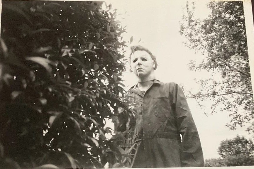 Michael Myers in a boiler suit and white mask, seen from below beside a dark bush and tall trees, in a black-and-white publicity photo.