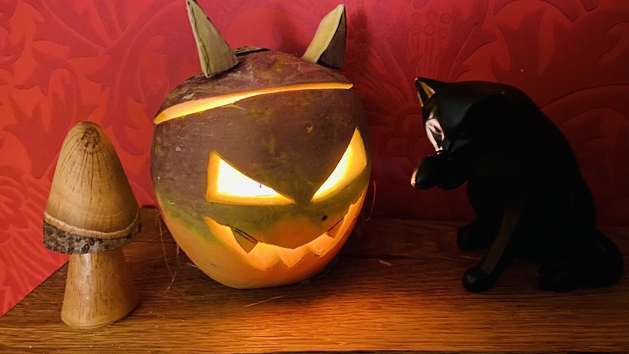 A glowing carved Scottish turnip lantern with an angry face and little ears, beside a black cat figure and a wooden toadstool.