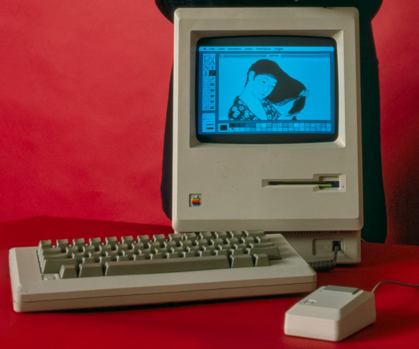 An original 1984 Apple Macintosh with its keyboard and one-button mouse, a black-and-white drawing on its small screen