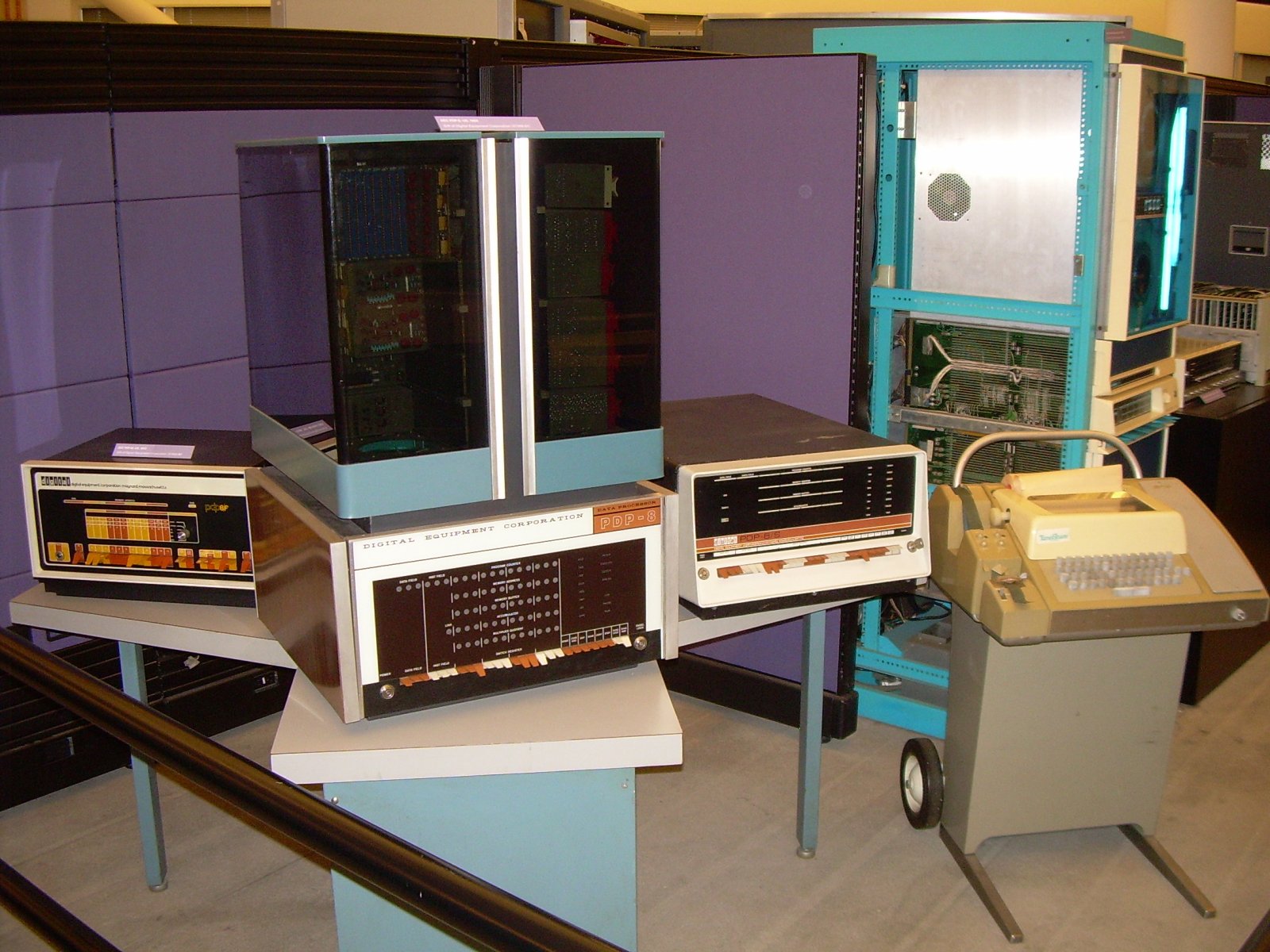 Vintage DEC minicomputers with rows of front-panel switches, beside a Teletype terminal, on display in a computer museum